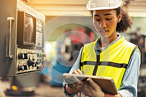 Standing in front of a control panel, a female industrial electrical engineer with a safety hardhat on her head and a tablet in