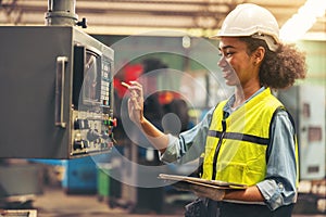 Standing in front of a control panel, a female industrial electrical engineer with a safety hardhat on her head and a tablet in