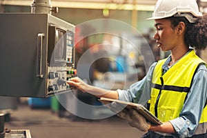 Standing in front of a control panel, a female industrial electrical engineer with a safety hardhat on her head and a tablet in