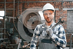 Standing in grey uniform and in white hard hat. Factory male worker is indoors