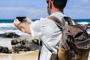 Standing man with backpack viewed from back take picture at the sea coast with smartphone device - concept of travel and summer