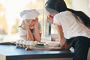 Standing and watching at each other. Mother with her daughter are preparing food on the kitchen