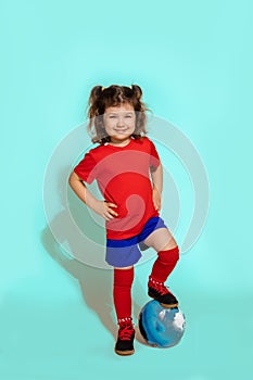 Photo of standing young soccer player girl 5-6 years holding football with foot on blue background, studio shot