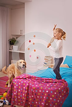 A star in the making. A little girl jumping on her bed and singing into her hairbrush while her dog watches.