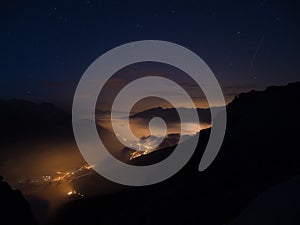 Starry sky and high mountain range, Italian French Alps, mist and fog in the vally with glowing villages below.