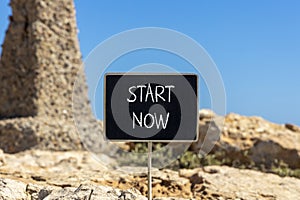 Start now symbol. Concept words Start now on beautiful black chalk blackboard. Beautiful brown stone blue sky background. Business