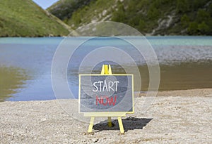 Start now symbol. Concept words Start now on beautiful black chalk blackboard. Beautiful mountain lake background. Business