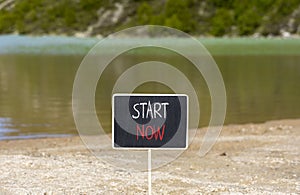 Start now symbol. Concept words Start now on beautiful black chalk blackboard. Beautiful mountain lake background. Business