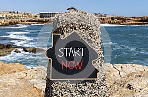 Start now symbol. Concept words Start now on beautiful black chalk blackboard. Beautiful stone sea blue sky background. Business