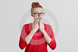 Startled frightened ginger woman covers mouth with hand, being afraid of something, wears spectacles and red sweater, poses alone