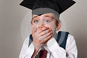 Startled schoolboy covered his mouth with hands. Boy in student hat. White background. Middle School