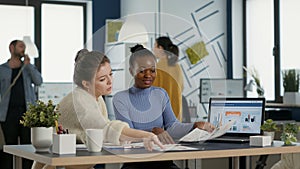 Startup employee working with african american coworker on group project comparing sales charts on clipboard