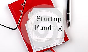 STARTUP FUNDING text on sticker on red notebook with pen and glasses