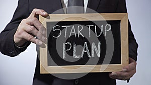 Startup plan written on blackboard, businessman holding sign, business concept