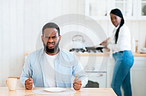 Starving Angry Black Man Waiting For Food At Table In Kitchen