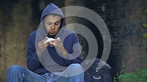 Starving troubled teen chewing snack sitting outdoors, dangerous city suburb