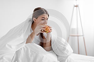 Starving upset hungry cute caucasian millennial woman sitting in bed at home and eating burger suffering from stress