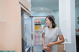 starving young asian woman hold her belly while open the fridge door looking for food