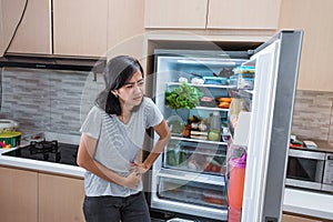 starving young asian woman hold her belly while open the fridge door looking for food