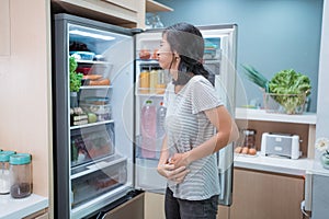 starving young asian woman hold her belly while open the fridge door looking for food