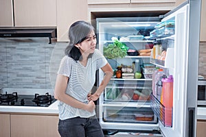 starving young asian woman hold her belly while open the fridge door looking for food