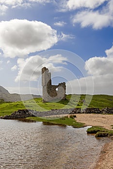 Stately Ardvreck ruins perched on green mound by serene lake, under vibrant cloudy sky