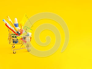 Stationery accessories in a shopping trolley on a yellow background with place for text