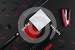 Pencils in black and red, stapler, red and black clips, ruler and knife, white paper on a black background