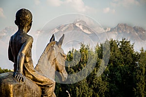 A statue of a boy riding a horse and viewing to snowy mountains slightly covered by clouds.