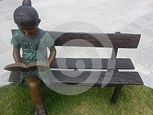 A statue of a girl reading a book while sitting on the wooden bench