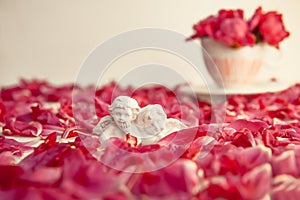 Statuette of two antique little lovely angels of the gypsum on the white background with red peony flower petals. Love, romantic,