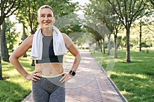 Stay active. Happy sporty woman in sportswear and wireless earphones smiling away, standing with towel around her neck