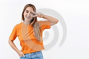 Always stay bright side. Portrait of optimistic happy blond girl in orange t-shirt, showing peace sign over eye and