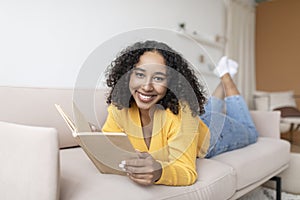 Stay home activities. Millennial black woman lying on sofa with open book, looking at camera indoors