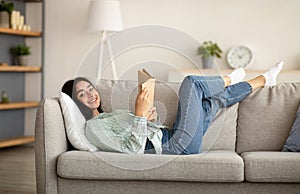 Stay home activities. Young Indian woman lying on couch with open book indoors, relaxing on Sunday morning