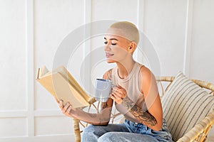 Stay home hobbies concept. Young lady reading book with cup of hot beverage, sitting in wicker chair in living room