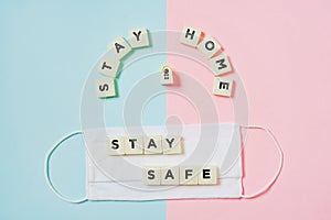 Stay home, plastic letter tiles,  with stay safe word on white fabric face mask on pink and blue paper background,