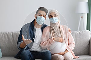 Stay At Home. Pregnant Muslim Couple In Medical Masks Sitting On Couch