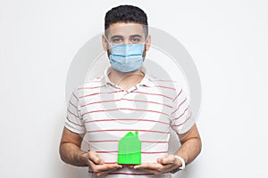 Stay at home, stay safe. Positive man with surgical medical mask holding green small house, looking at camera kindly, reassuring