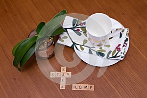 Stay Safe Home message written on walnut table set with white plate, napkin, coffee cup and small houseplant
