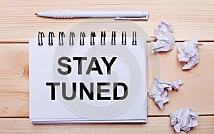 STAY TUNED written in a notebook near a white pen and pieces of paper on a wooden background