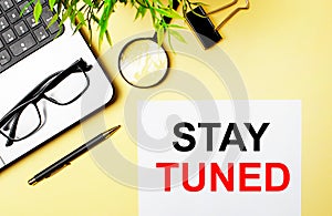 STAY TUNED is written in red on a white piece of paper on a light yellow background next to a laptop, pen, magnifying glass,