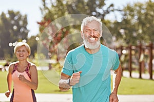 Staying healthy and fit. Happy mature man smiling at camera while running together with his wife in the early morning