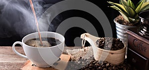 Steam coffee cup with grinder,roasted beans and flower pot on wood background in morning sunlight