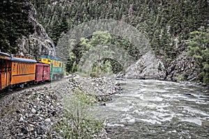 Steam engine rounding a curve with mountain cliff on one side and rushing river on other in pine forested mountains - Vintage