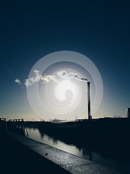 Steam rises from a smokestack on a factory silhouetted against the sunlight.