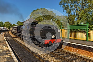 A steam train going past a station towards a single track section of line on a railway in the UK