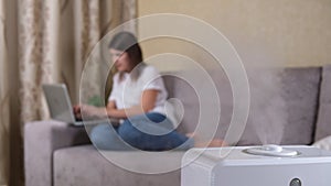 Steam from a working humidifier with ionization of air in the room, against a blurred background, woman working at laptop sitting