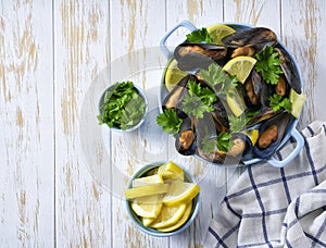 Steamed Mussels in shells in a blue plate with herbs and lemon. White wooden background, top view