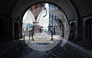 Steampunk style city streets with a view through a tunnel and a airship steam powered hot air balloon in the background .